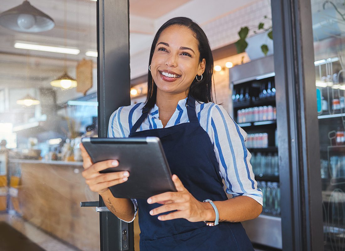 Business Insurance - Closeup Portrait of a Smiling Young Business Woman Wearing an Apron Standing Outside her Small Cafe Shop While Holding a Tablet