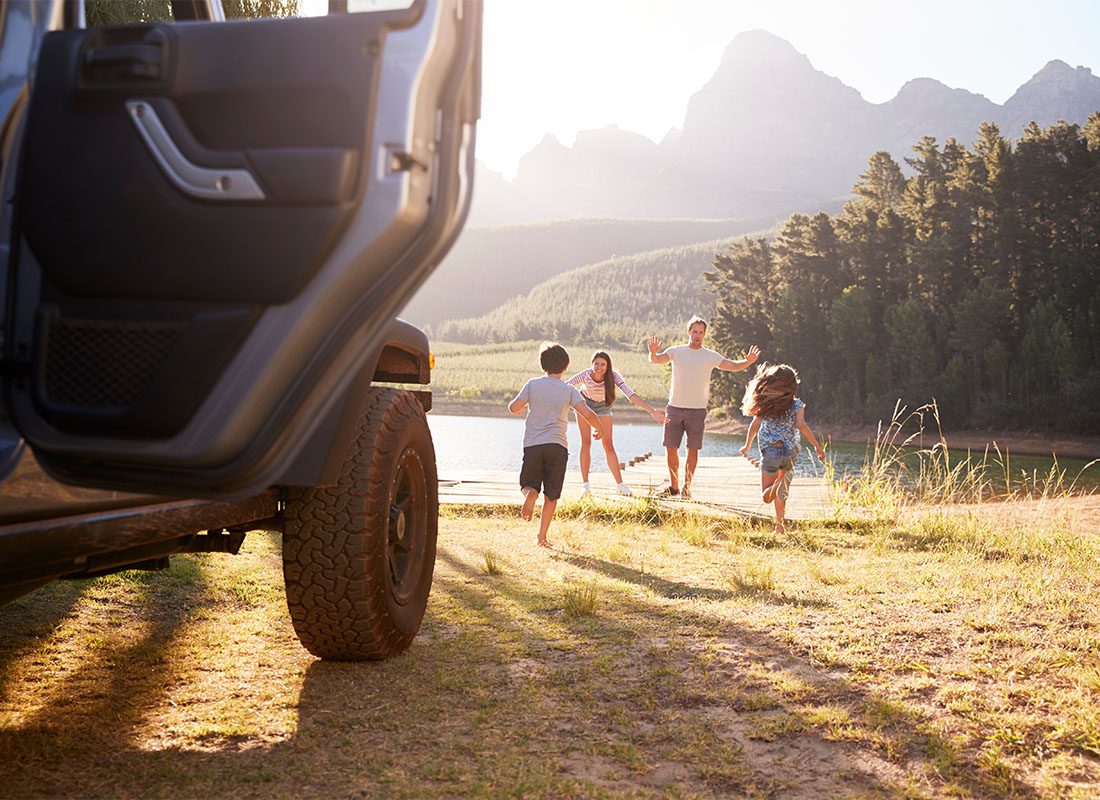 Insurance Solutions - View of a Car with the Back Door Open and Two Kids Running to Their Parents Standing Next to the Lake During a Summer Road Trip